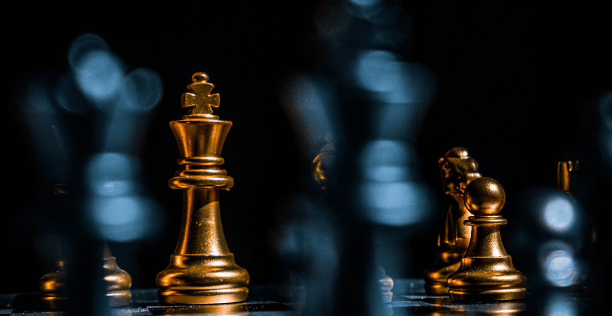 Um tabuleiro de xadrez onde as peças douradas representam as estratégias divinas e as peças acinzentadas as movimentações humanas.
